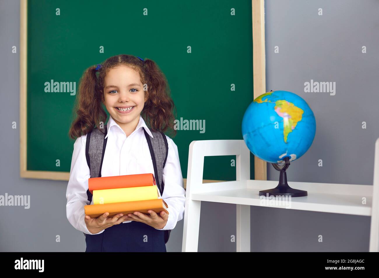 Smiling girl with backpack holding pile of books near blackboard at classroom. Cute pupil happy to go back to school Stock Photo