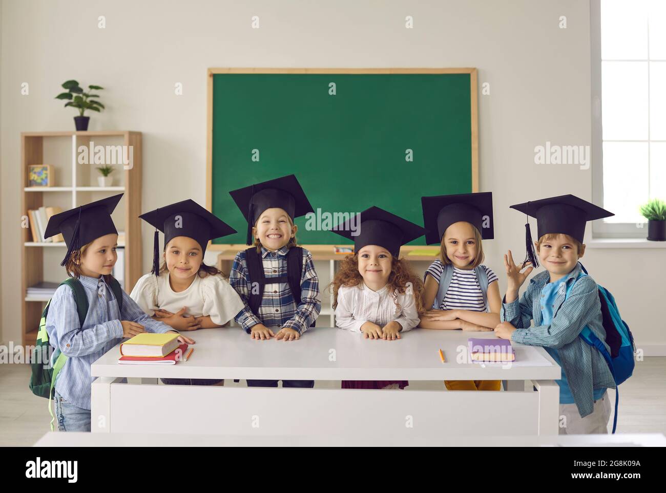 Group of happy school kids in academic caps standing by table with books in classroom Stock Photo