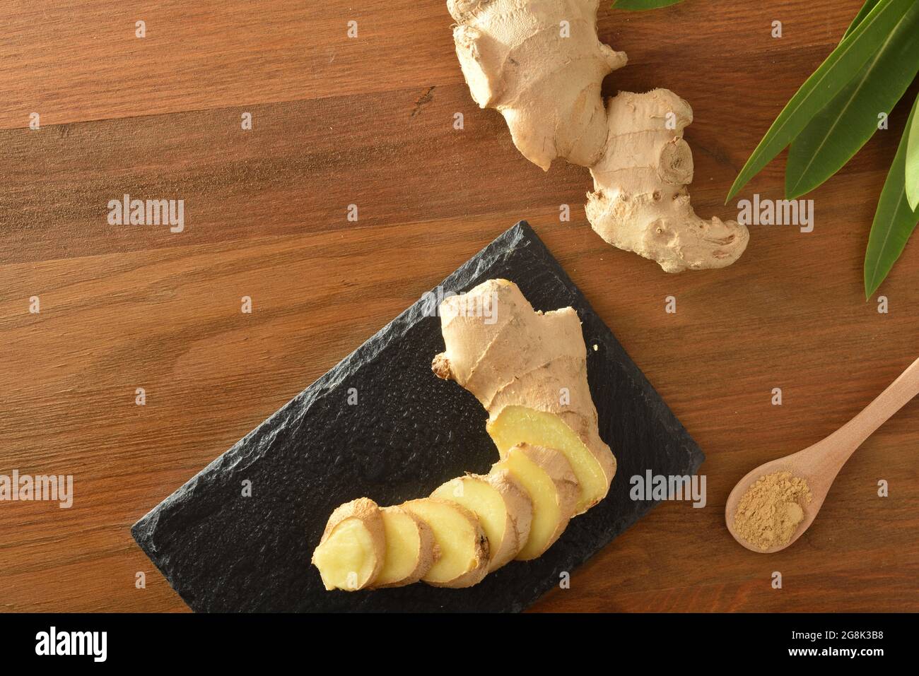 Ginger root slice on slate plate and spoon with powder on wooden table with root and leaves. Top view. Stock Photo