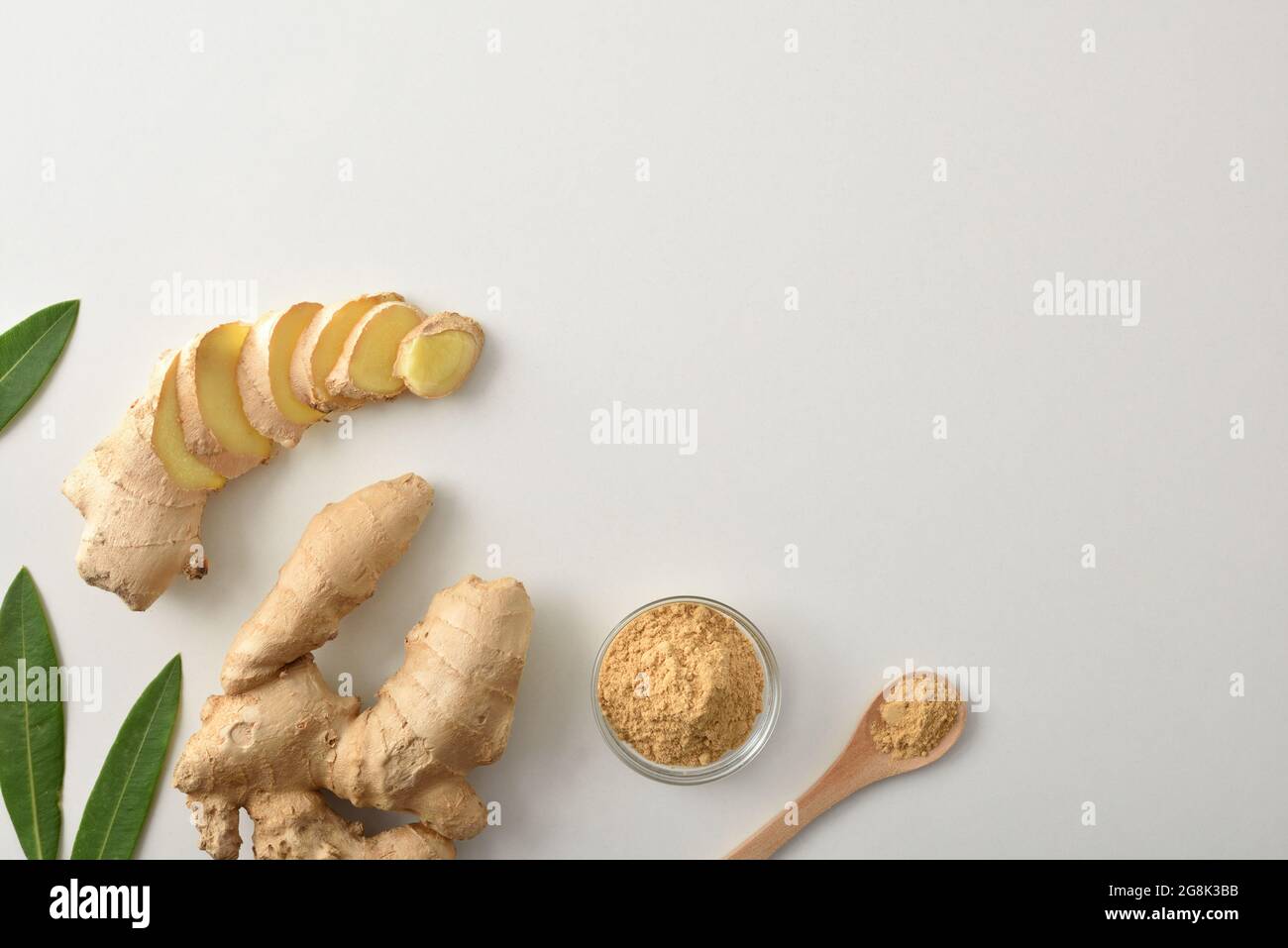 Ginger root and with slices and powder in glass jar and wooden spoon on white table with leaves. Top view. Stock Photo