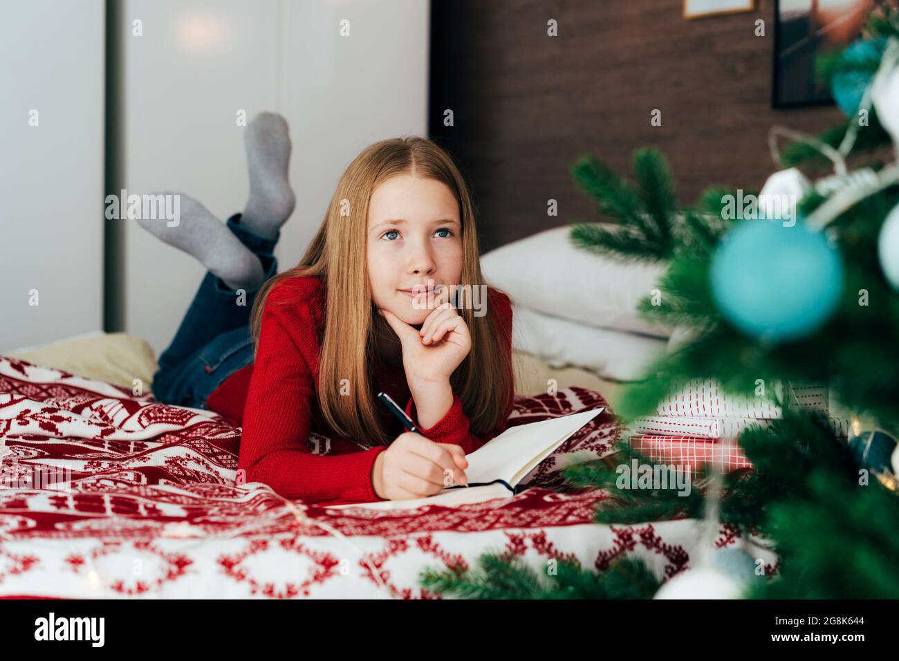 Red-haired caucasian teenager girl lying on the bed writing a letter to Santa Claus. Stock Photo