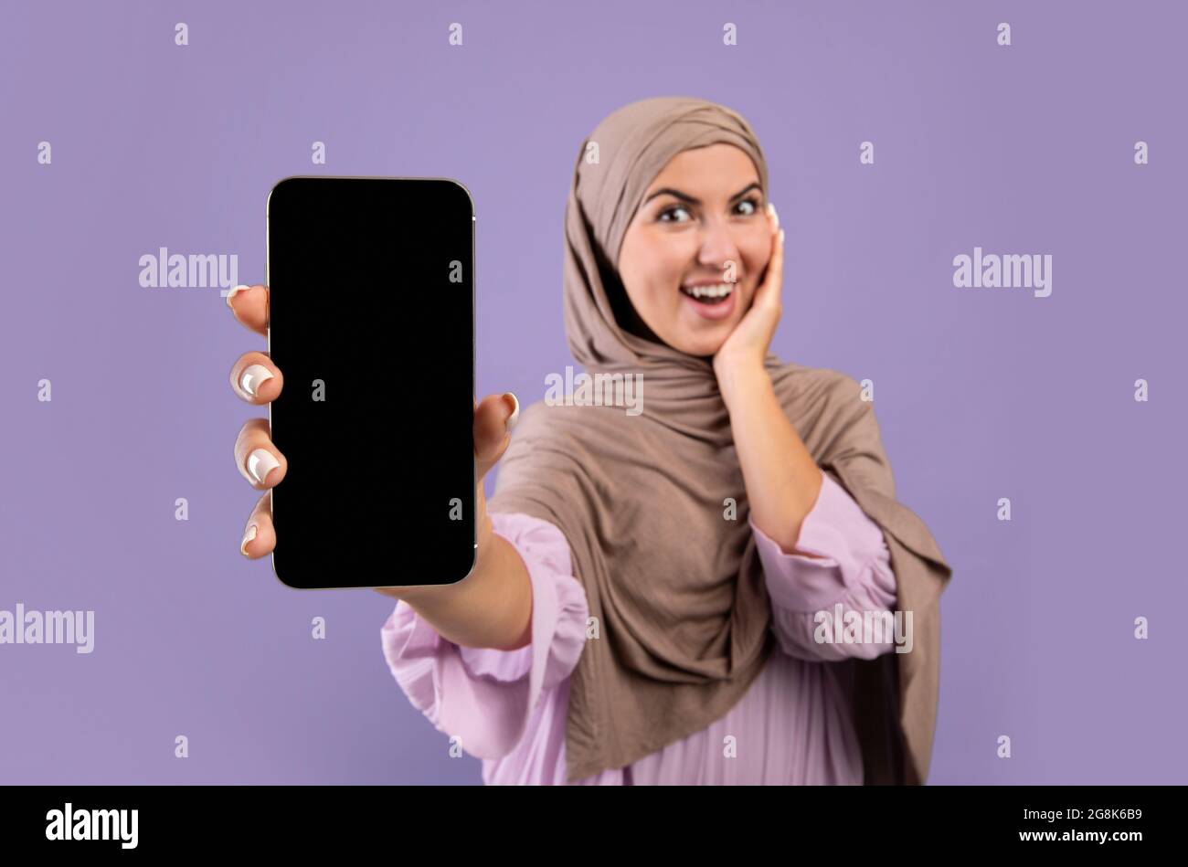 Mockup image of smartphone with black blank screen in hands of happy muslim woman in hijab over purple background Stock Photo