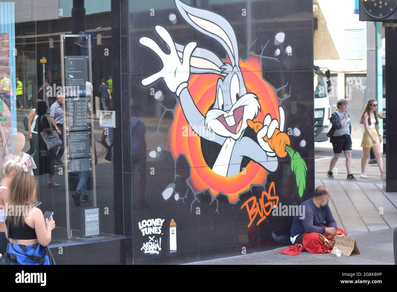 Manchester, UK. 21st July, 2021. UK Weather: Another glorious, sunny afternoon in Manchester, England, UK, as the heatwave continues. A man with a card sign saying he is 'very hungry god bless' sits by a cartoon figure on the wall, while people pass by. The cartoon figure is a Bugs Bunny image, part of a Looney Tunes art trail, in the city centre. Credit: Terry Waller/Alamy Live News Stock Photo