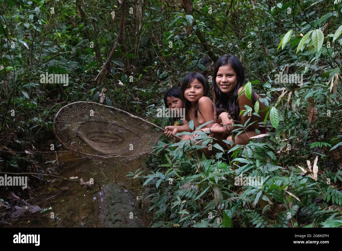 Three girls stood with a fishing net next to the river. AMAZONIAN ECUADOR: In one image, a woman wore red makeup across her eyes and a feathered headd Stock Photo