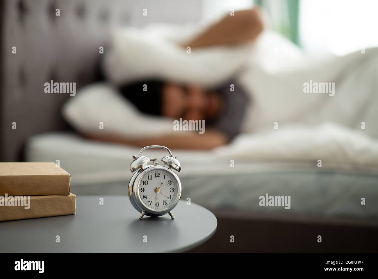 Bad sleep concept. Exhausted eastern man being awaken with alarm clock in his bedroom, covering ears with pillow Stock Photo