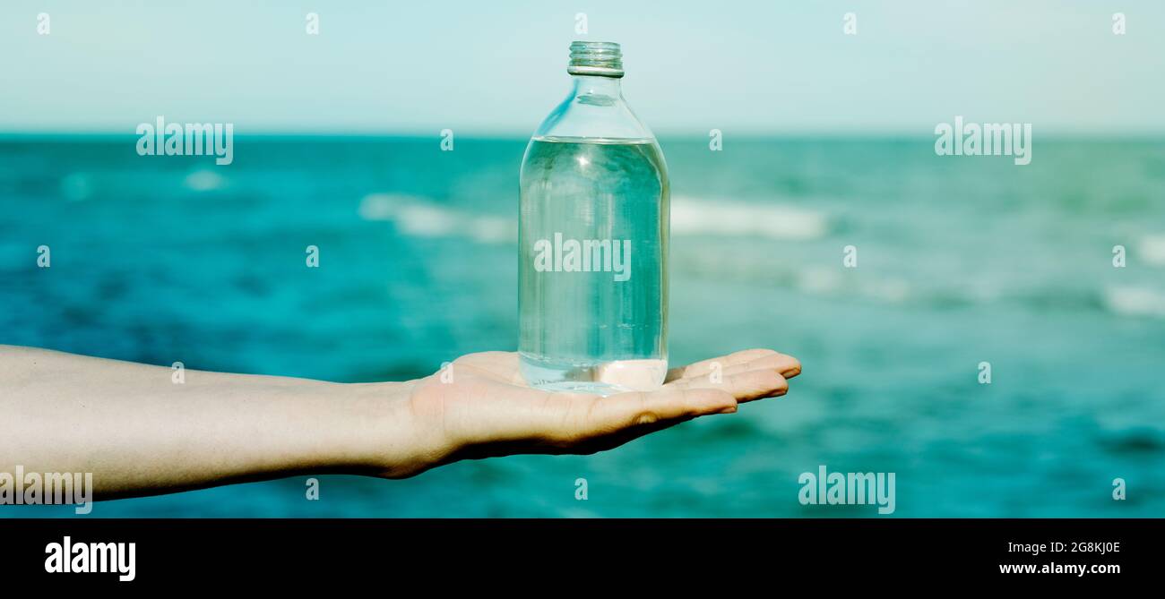 closeup of a young caucasian man holding a glass reusable bottle full of water in the palm of his hand, in front of the ocean, in a panoramic format t Stock Photo