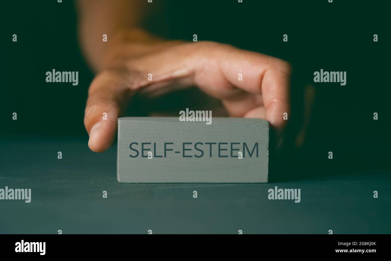 closeup of a caucasian man showing a gray building block with the text self-esteem written in it, on a dark gray surface Stock Photo