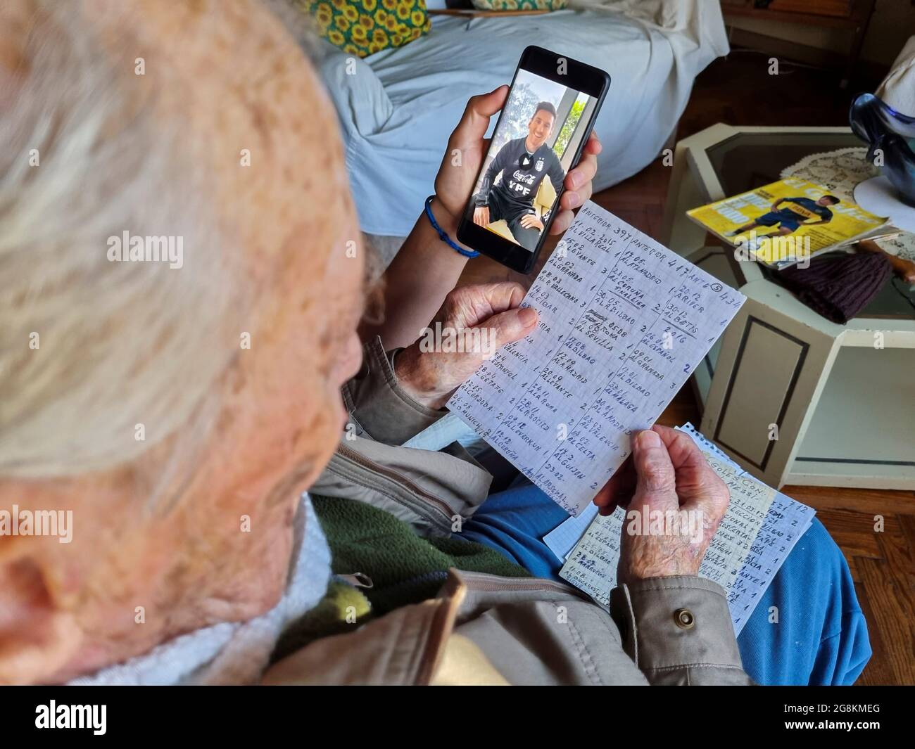 Hernan Mastrangelo, a 100-year-old former chocolatier and a fan of Argentine soccer star Lionel Messi, holds his handwritten notes of Messi's goals, as his grandson Julian holds a mobile phone with a personalised message from Messi, in Buenos Aires, Argentina July 20, 2021. Picture taken July 20, 2021. REUTERS/Miguel Lo Bianco Stock Photo