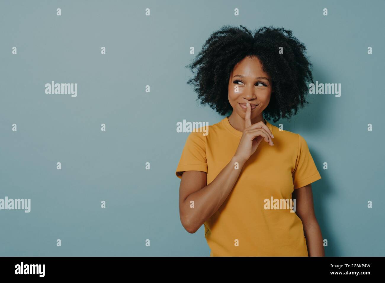 Cheerful afro girl showing shhh sign with finger near lips, standing over pastel blue background Stock Photo