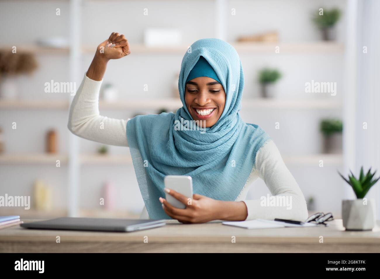 Overjoyed black muslim woman in hijab looking at smartphone and celebrating success Stock Photo