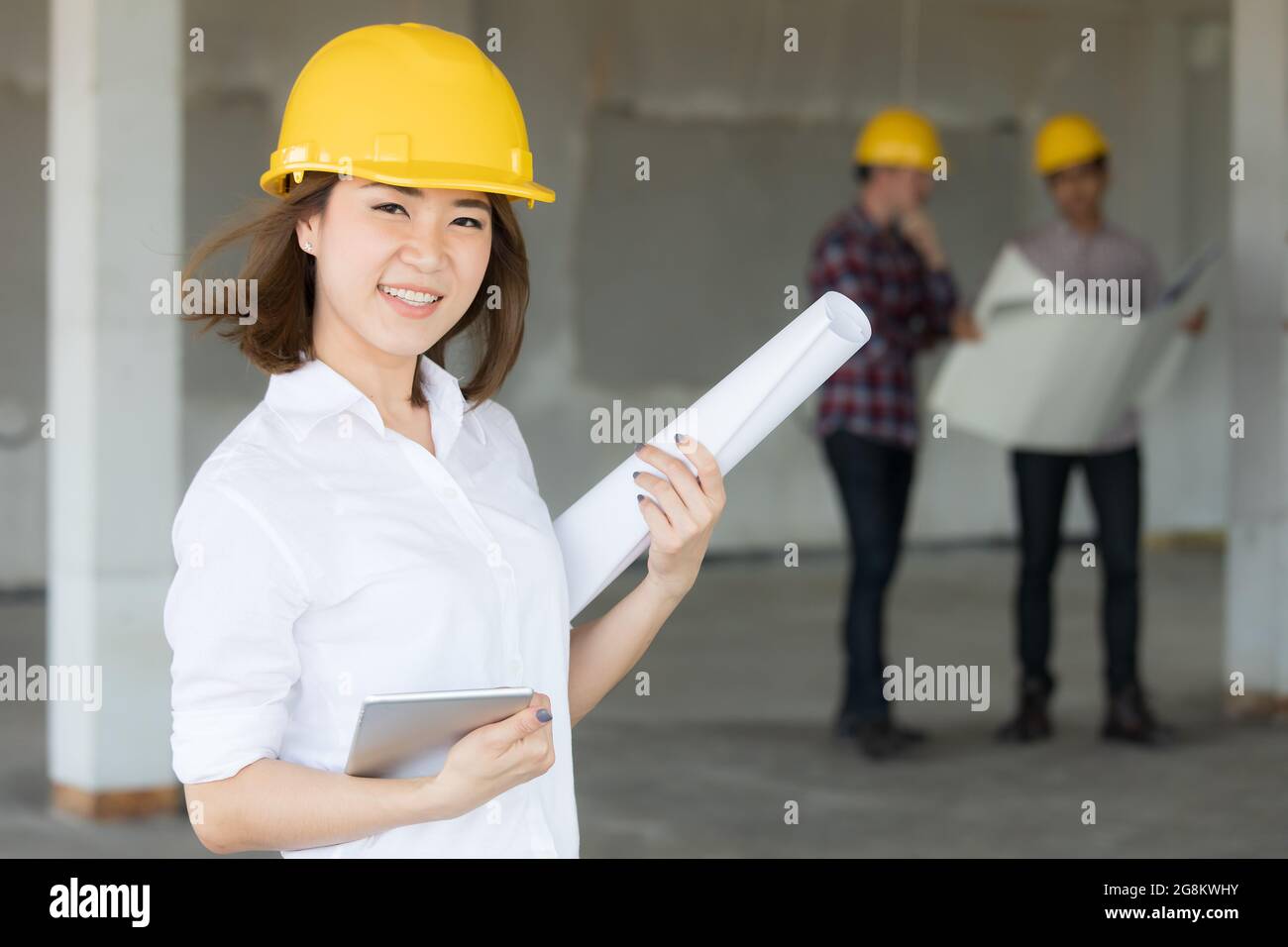 Female Asian enginee holding blueprint plan and tablet computer in hands standing and smiling with selt confident while two male team talking in blur Stock Photo