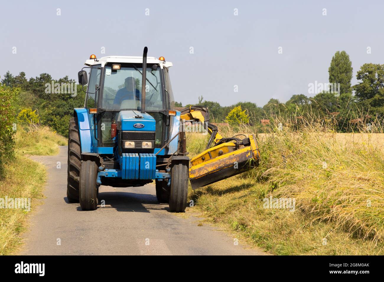 Man in a tractor mowing grass verges in a country lane in summer. Much Hadham, Hertfordshire July 2021 Stock Photo