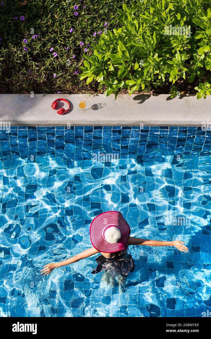 Woman wear big hat spread arms and walking in swimming pool going to glass of orange juice and red headphone. Stock Photo