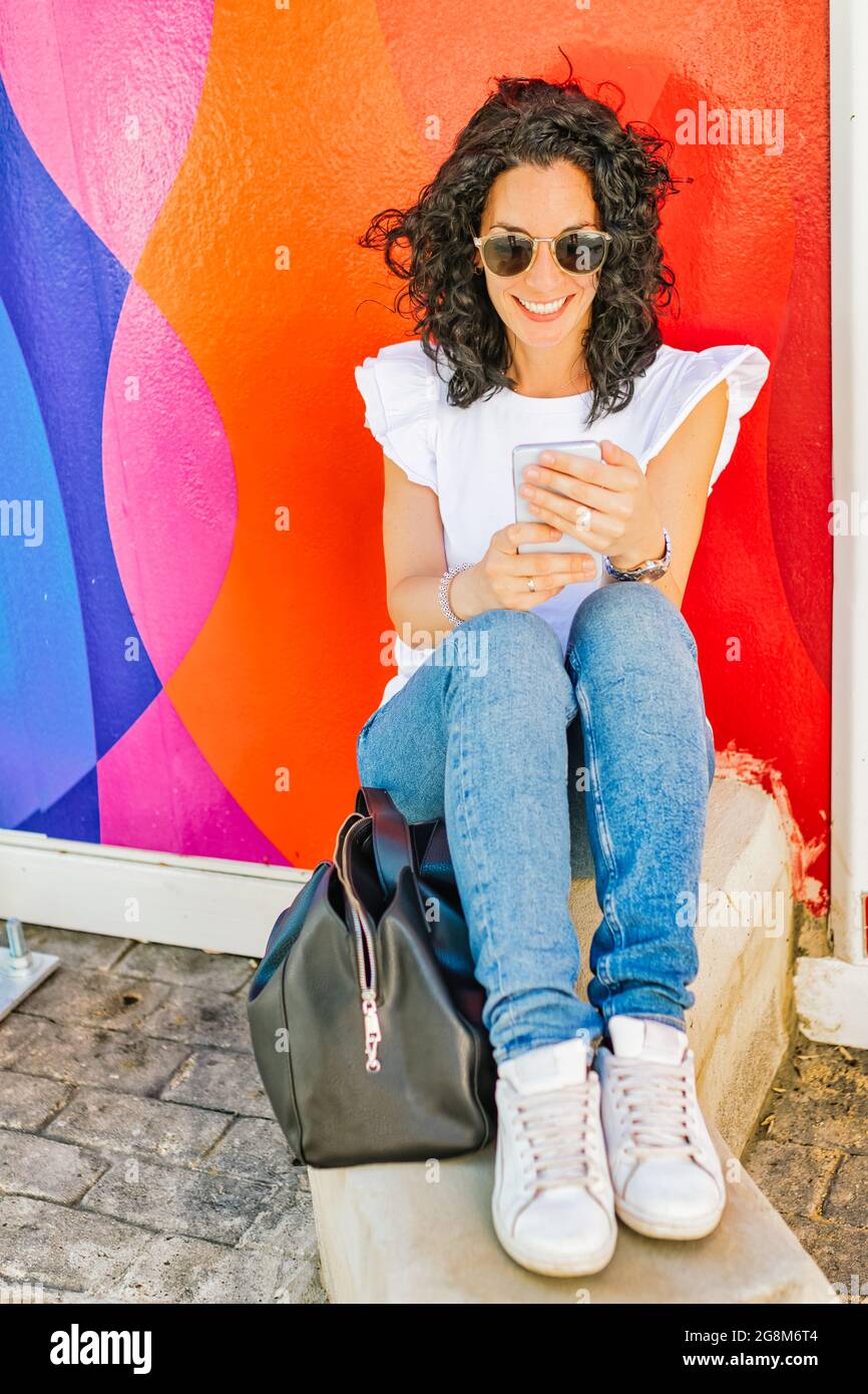young woman with curly black hair has fun chatting with her mobile sitting on a colorful wall in the street. Stock Photo