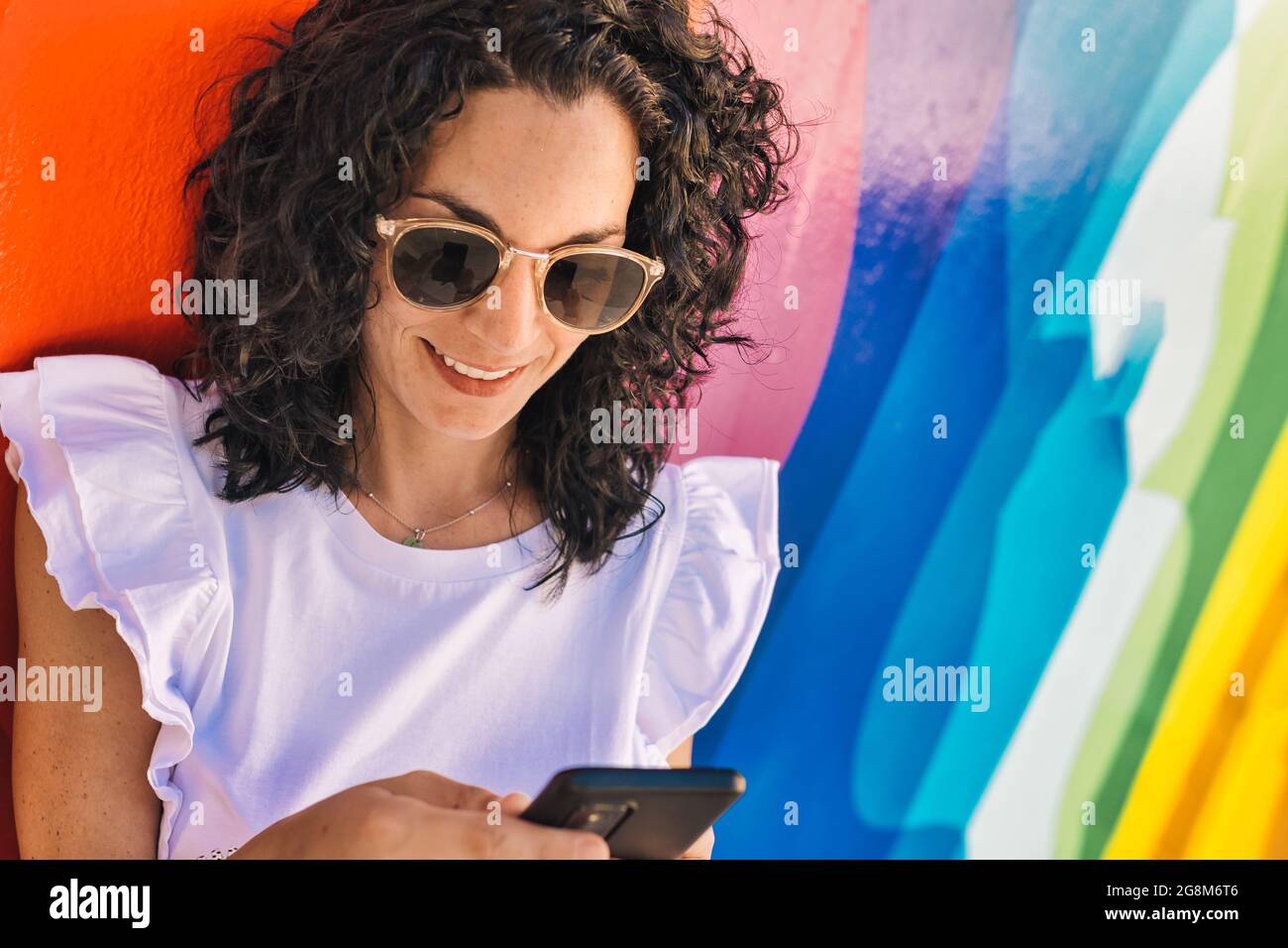 young woman with curly black hair has fun chatting with her mobile sitting on a colorful wall in the street. Stock Photo