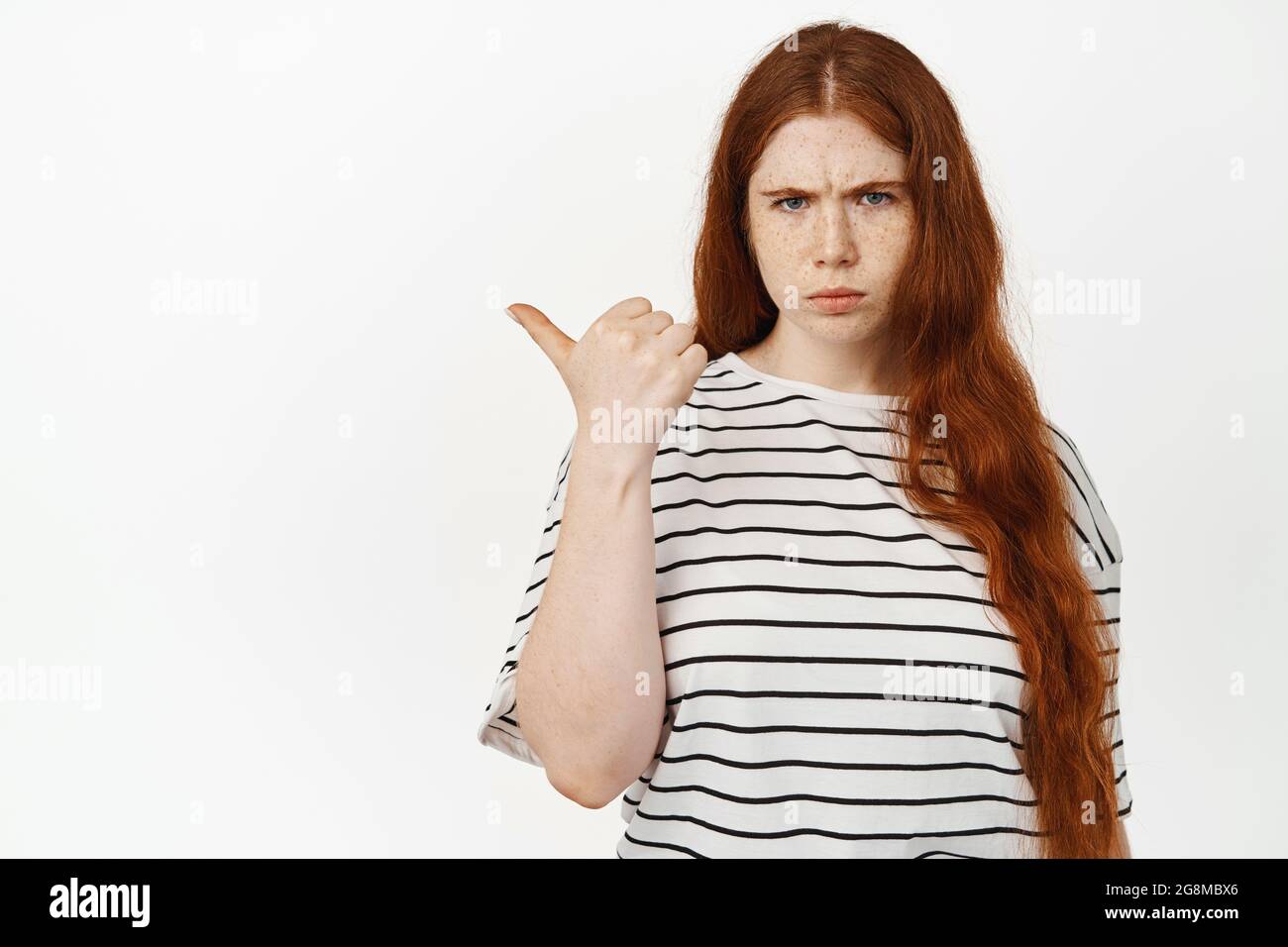 Image of angry and upset teen girl, redhead female student furrow eyebrows, looking with frustrated face and pointing finger left, showing smth with Stock Photo