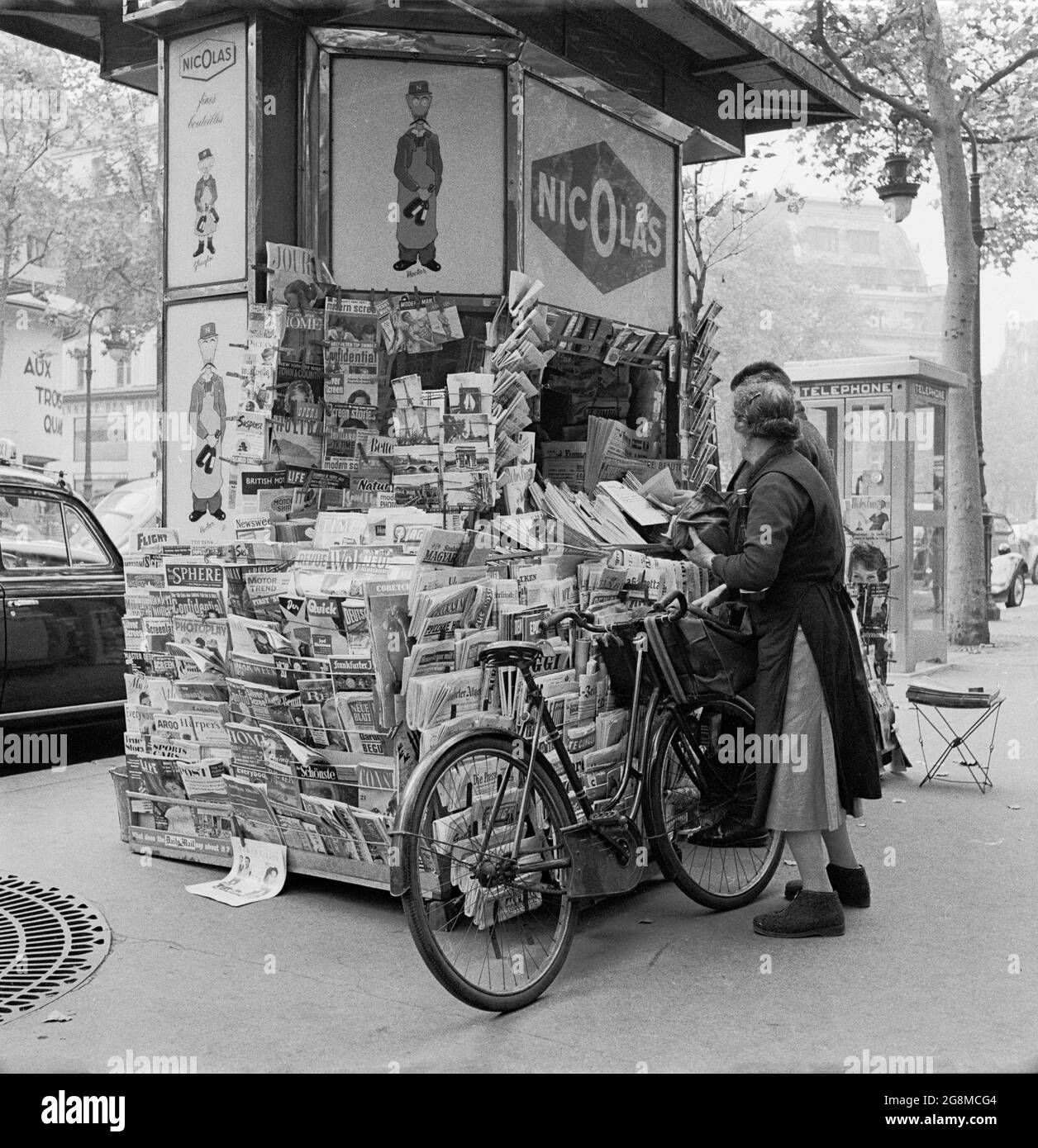 1950s, historical picture of two people, one a lady with her bicycle outside at a newspaper kiosk in central Paris, France. They first made an appearance in 1857, when Baron Haussmann was making renovations to the Parisian landscape. The kiosk is full of newspapers, the reason being that under French law, all the newspaper titles had to be carried, so all political opinion was on display. On the kiosk is advertsing for Nicolas, the long established parisian wine company. Stock Photo