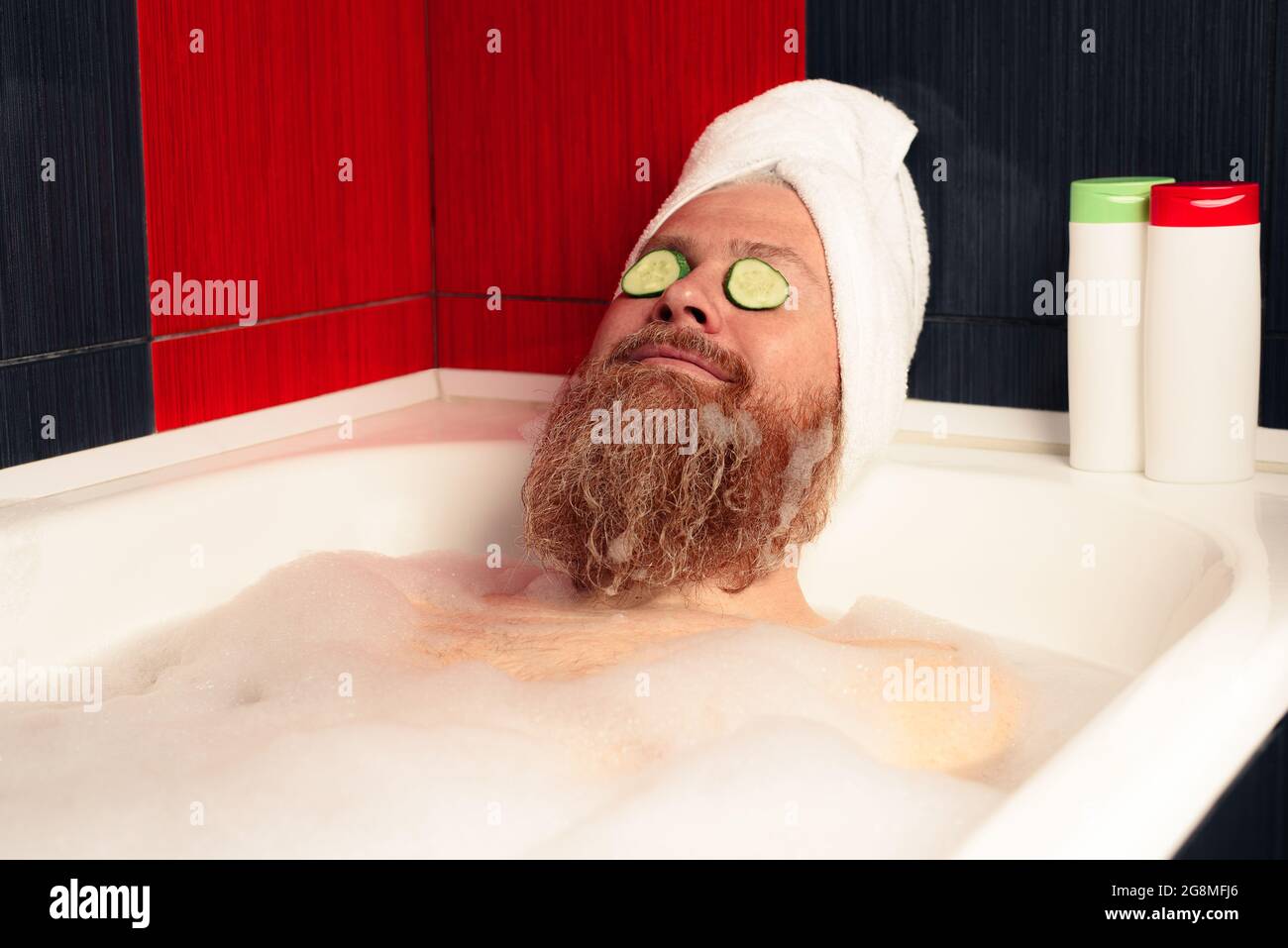 Cute bearded man taking bath with head wrapped in towel and cucumber slices on his eyes. Funny hipster relaxing in foamy bathtub and enjoying life Stock Photo