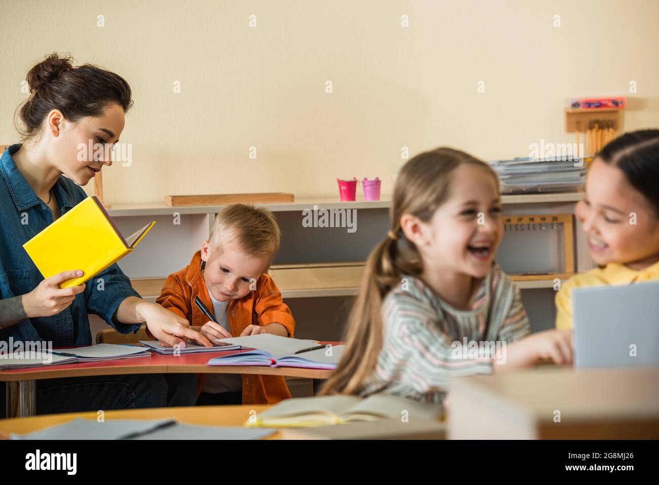 teacher pointing at notebook near writing boy while interracial girls laughing on blurred foreground Stock Photo