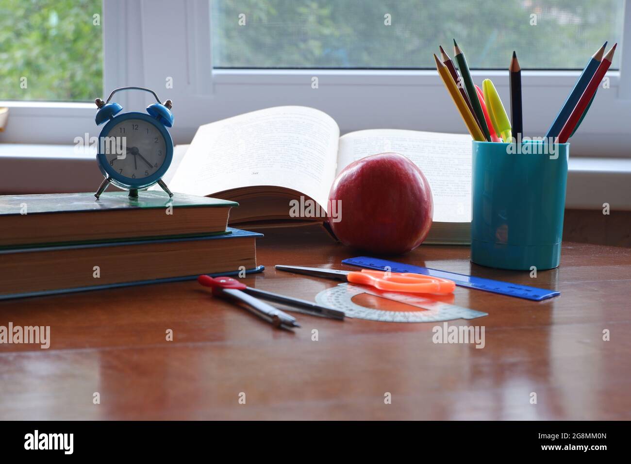 Opened book on the table near the window with an apple and an alarm clock. Stock Photo