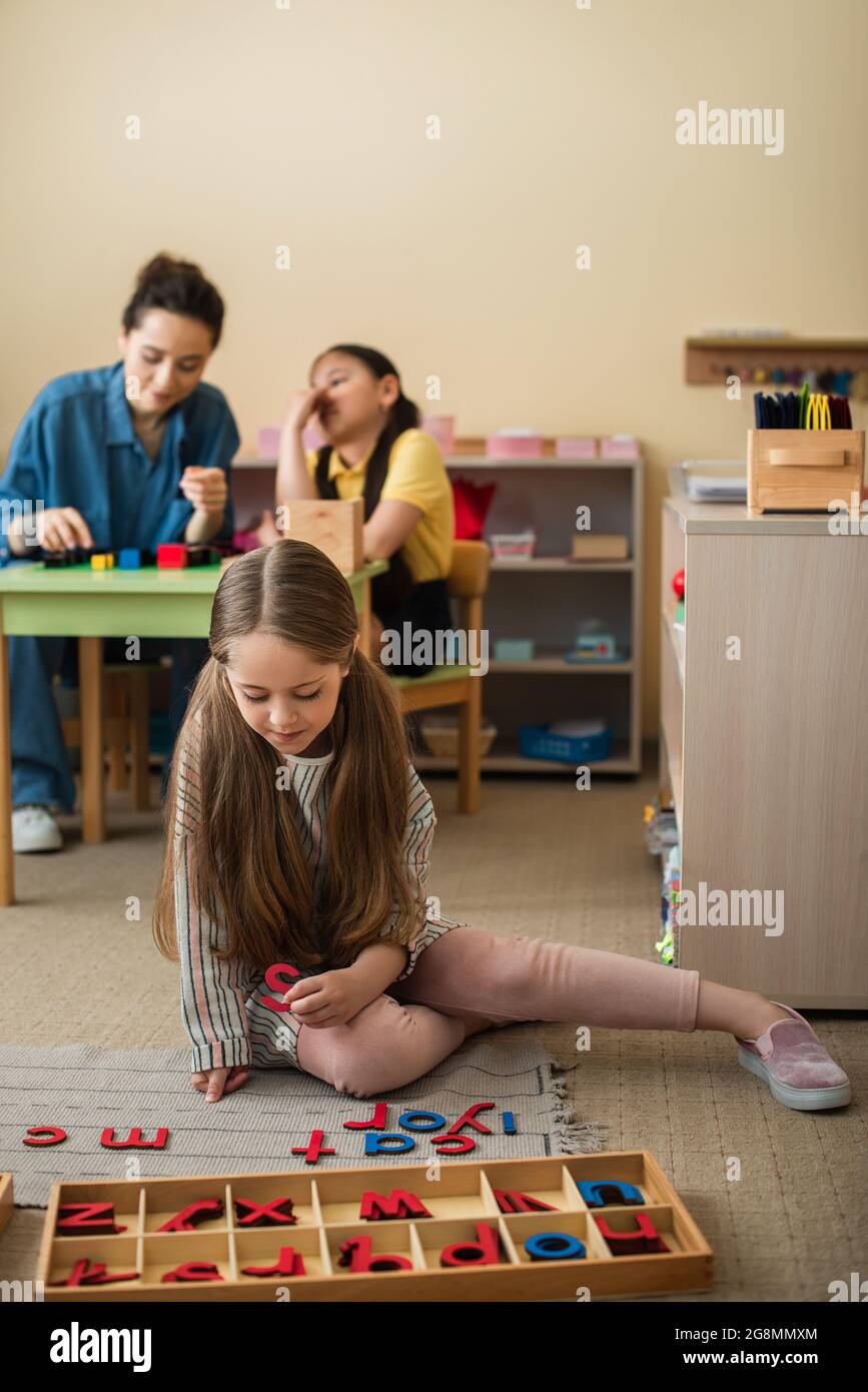 child on floor near wooden letters and teacher playing game with asian girl on blurred background Stock Photo