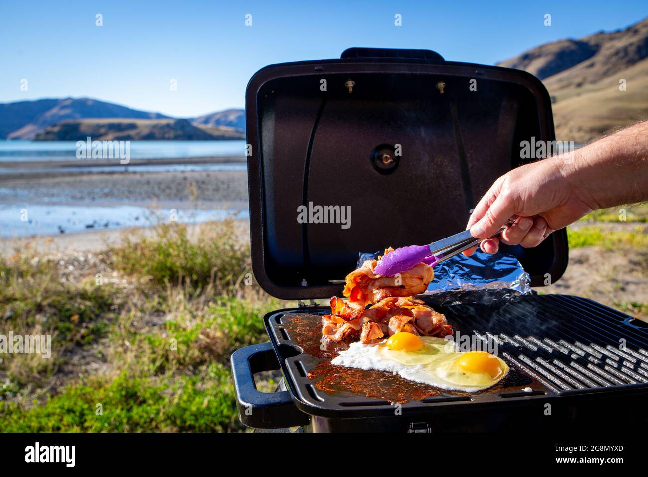 A man cooks bacon and eggs on a portable bbq beside the seaside on a sunny winter day travelling in New Zealand Stock Photo