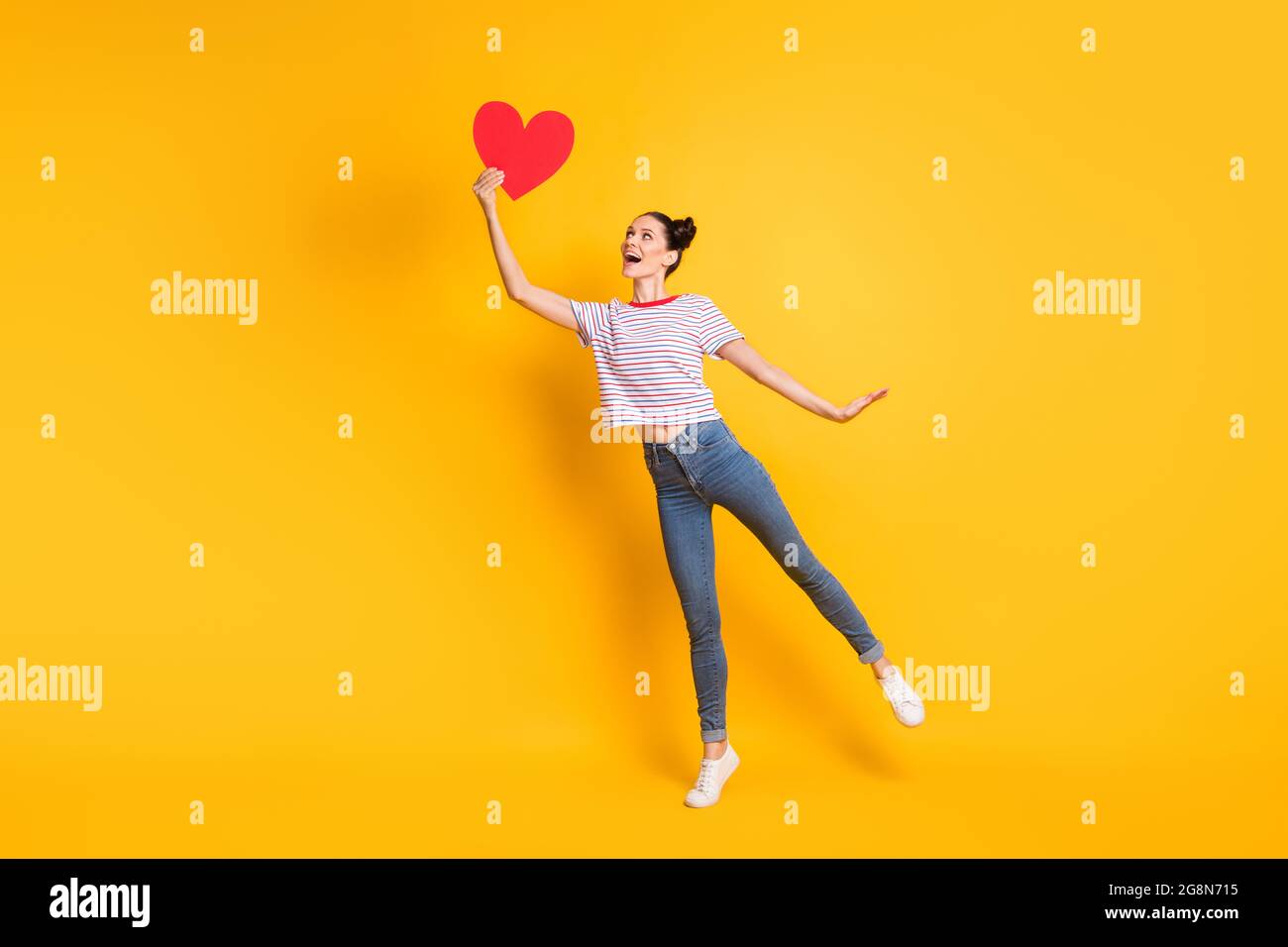 Photo of sweet adorable young girl dressed striped t-shirt catching red paper heart isolated yellow color background Stock Photo