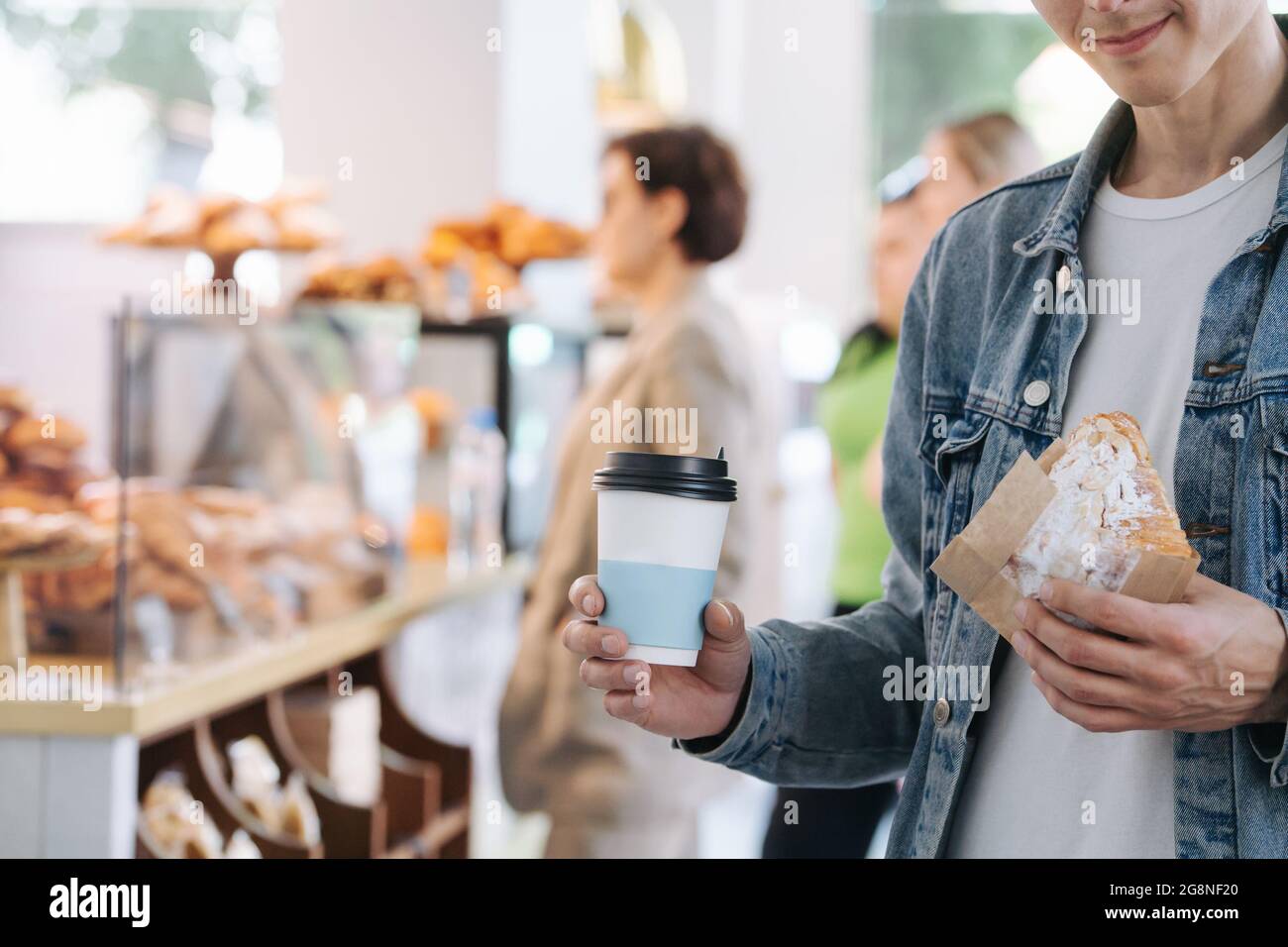 Bokeh image of a man in a jeans jacket holding cup of coffee and croissant in a paper wrap. Cropped, half face, smiling. Stock Photo
