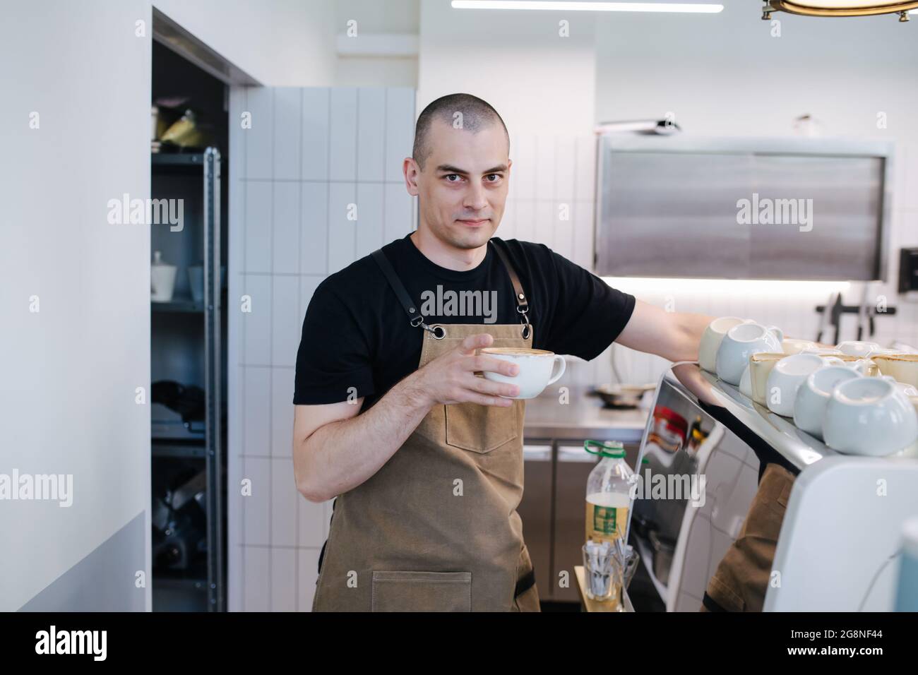 Portrait of a bold male baker posing with coffee cup in hands. Inside a disorderly busy bakery kitchen. Looking at the camera. Stock Photo