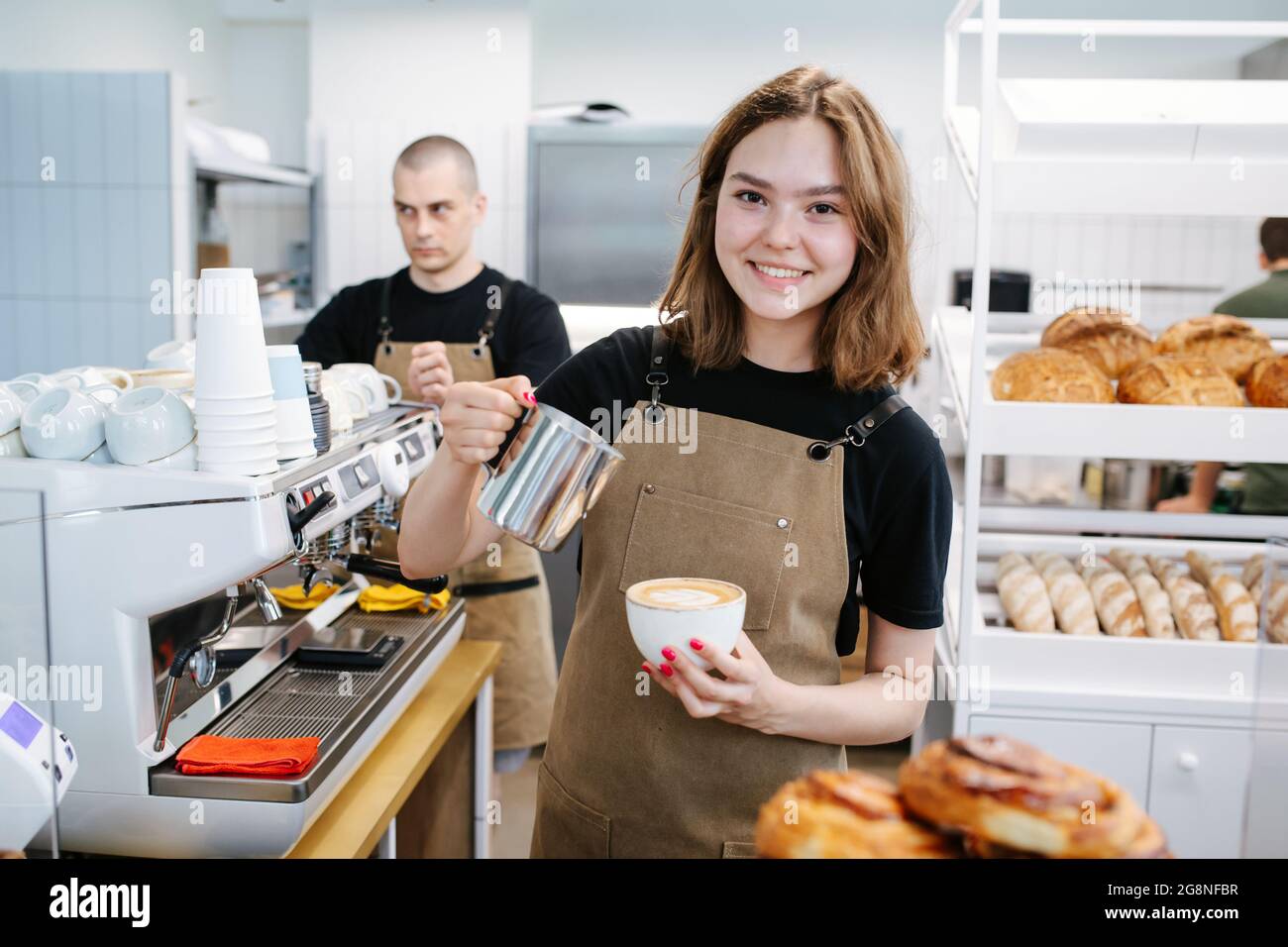 Young beautiful baker girl posing with coffee and cream jug in hands. Charmingly smiling at the camera. Inside a disorderly busy bakery kitchen. Stock Photo