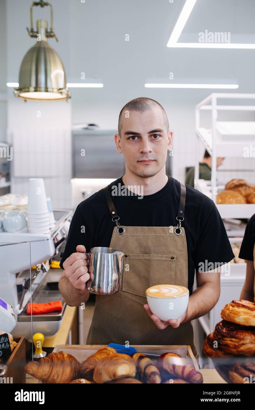 Handsome bold male baker posing with coffee and cream jug in hands. Inside a disorderly busy bakery kitchen. Looking at the camera. Stock Photo