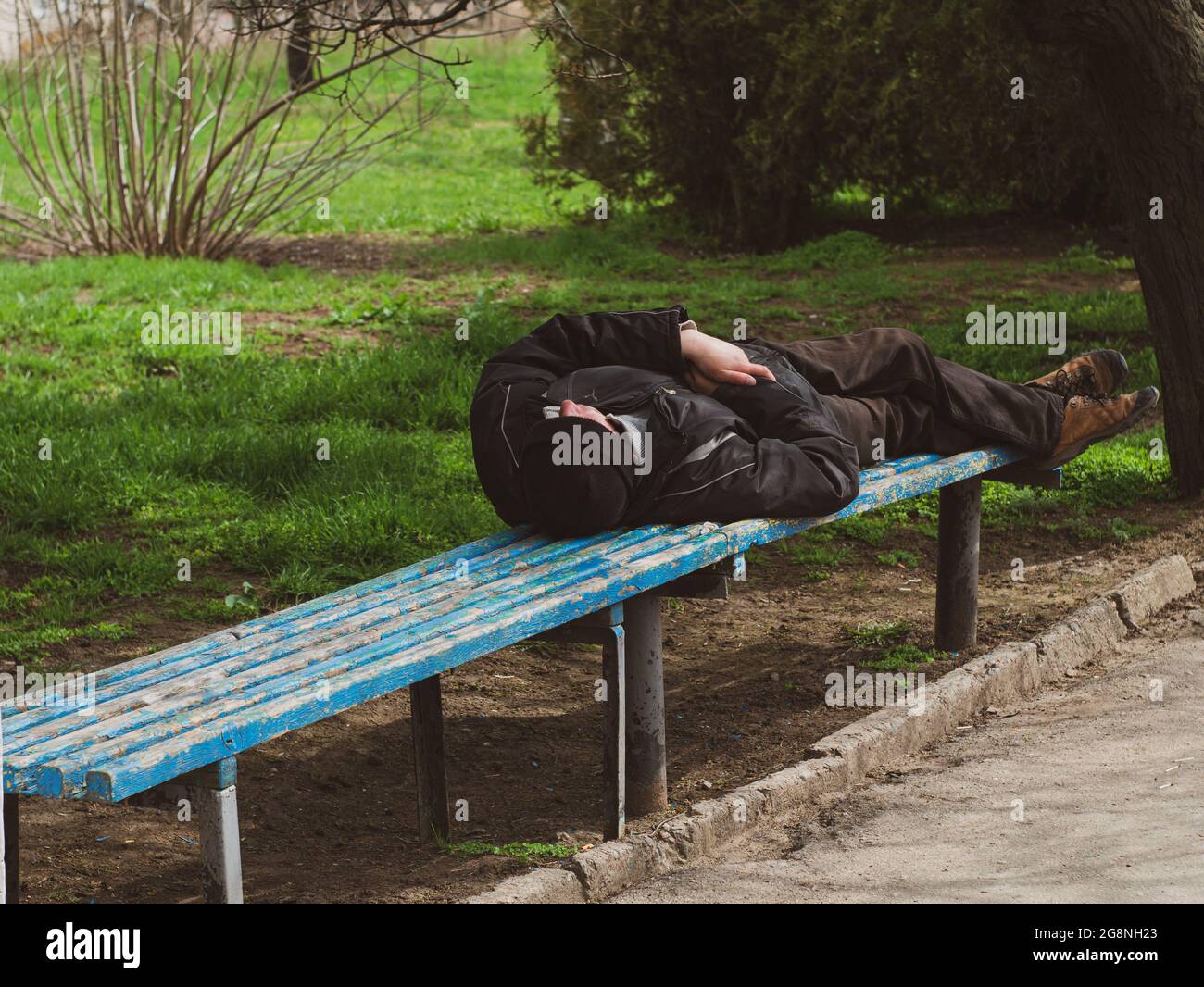 Sleeping homeless man on bench in the park with green grass. Drunk vagabond. Berdiansk, Ukraine - 18 april 2021 Stock Photo