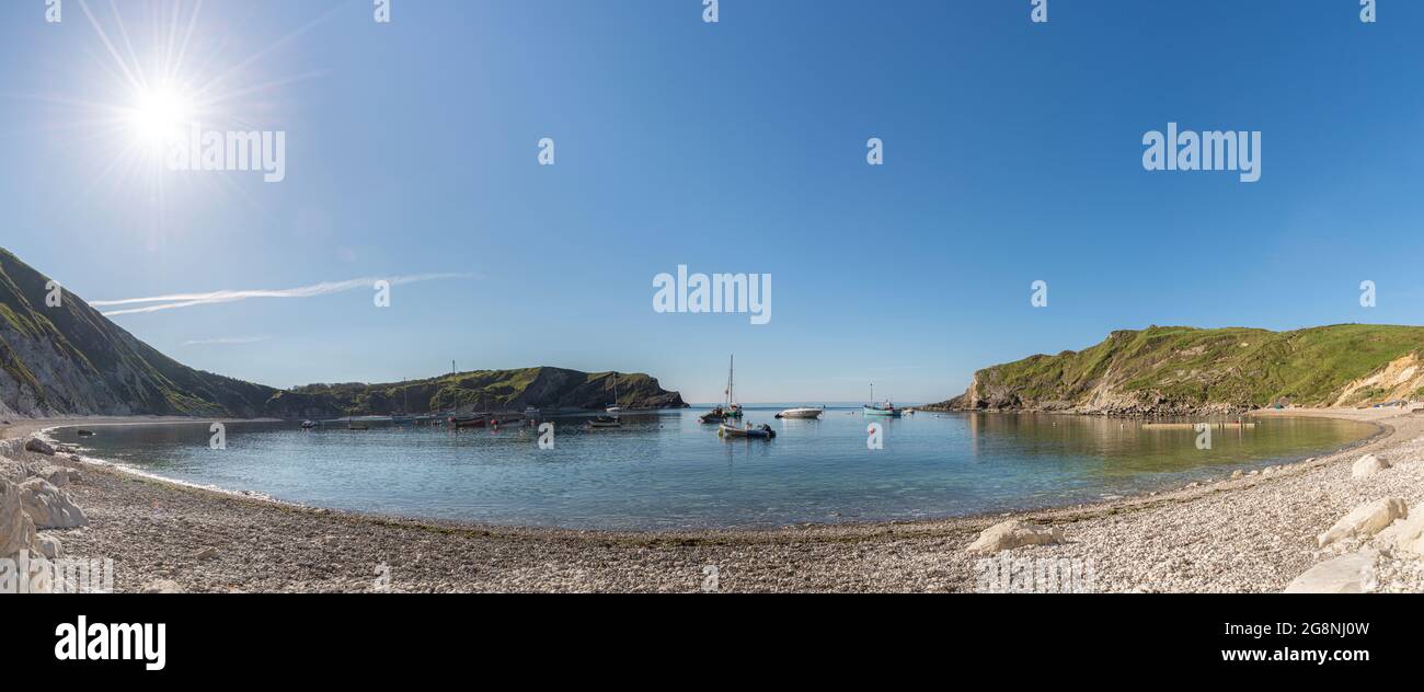 Far side of Lulworth cove with the morning sun lighting up the pebbles and rocks on the beach, with sailing boats anchored in the cove, West Lulworth, Stock Photo