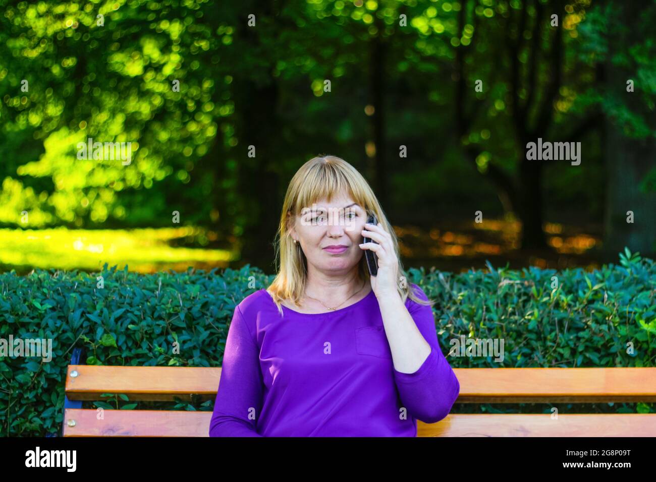 Defocus caucasian blond woman talking, speaking on the phone outside, outdoor. 40s years old woman in purple blouse in park on bench. Adult female peo Stock Photo