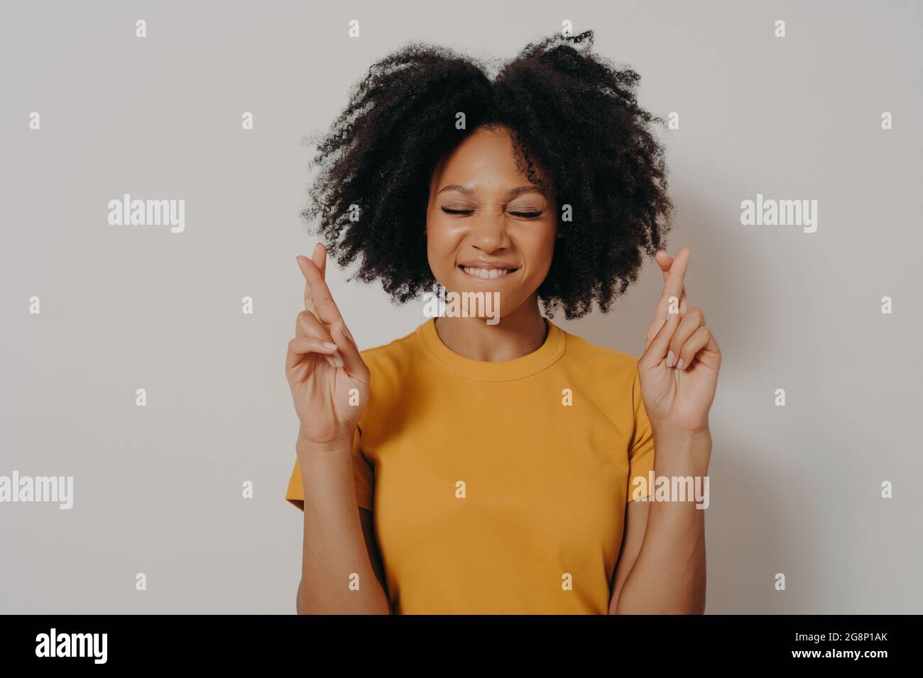 Portrait of pretty cheerful african girl standing in studio and holding fingers crossed for luck Stock Photo