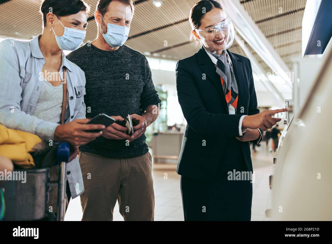 Ground staff helping couple with self service check-in during pandemic. Man and woman in face masks helped by airlines attendant at airport in pandemi Stock Photo