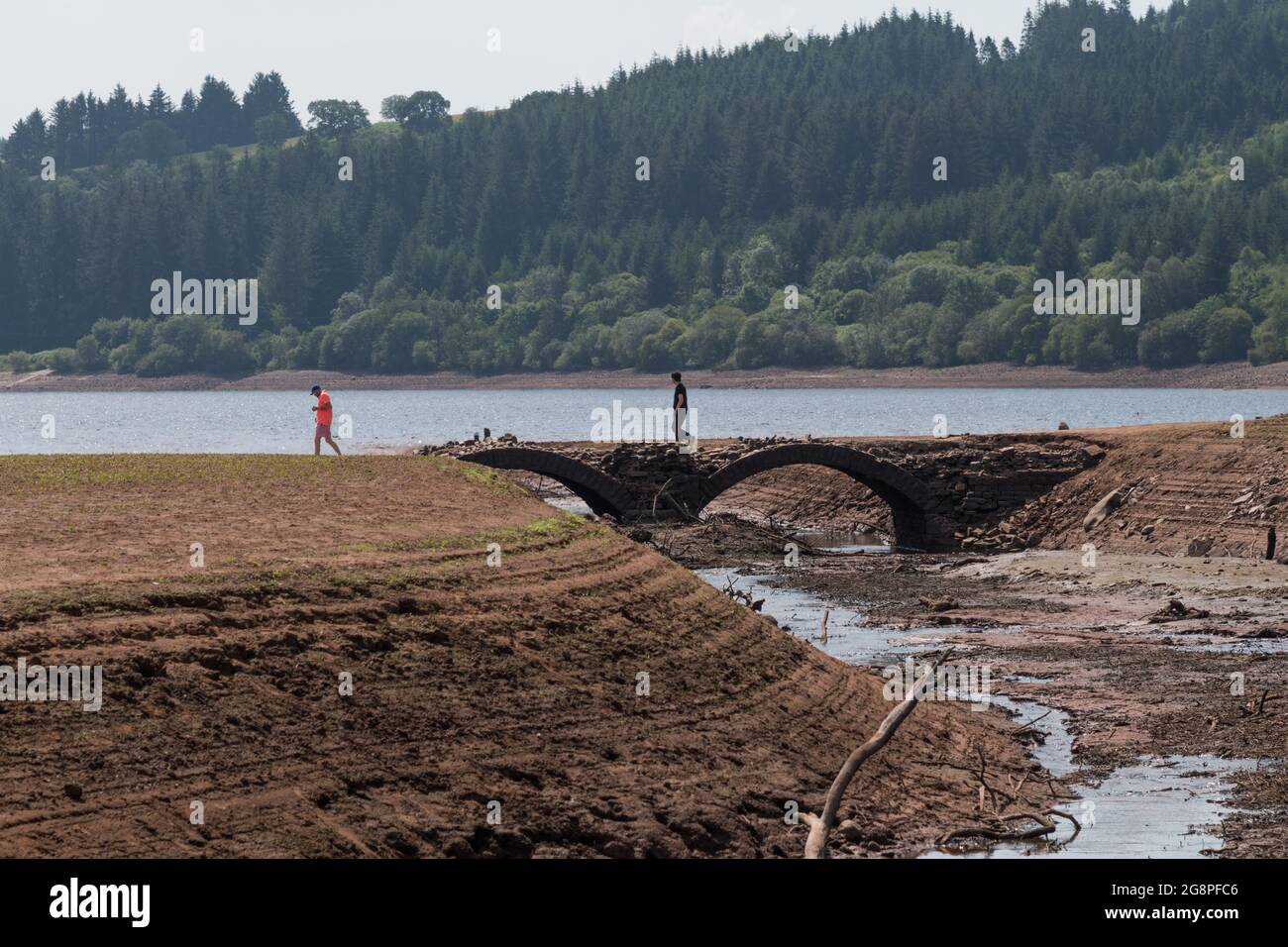 Llwyn Onn reservoir, Merthyr Tydfil, South Wales, UK.  22 July 2021.  UK weather:  Water levels continue to drop at this reservoir as the heatwave continues. People walk across an old bridge normally underwater, uncovered by the low levels.  Credit: Andrew Bartlett/Alamy Live News Stock Photo