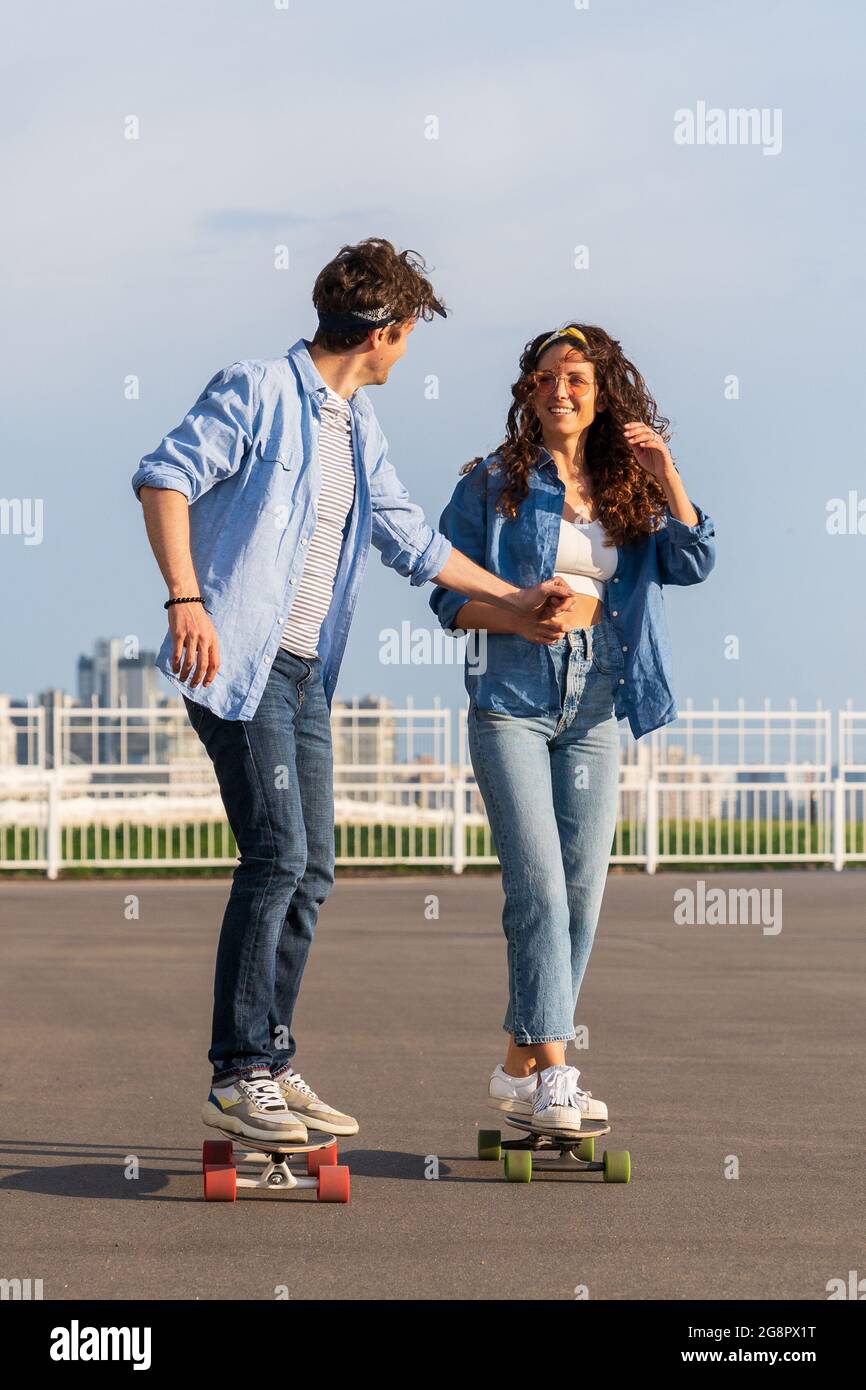 Stylish couple skateboarding together over urban skyline. Young adult man and woman on londboard Stock Photo