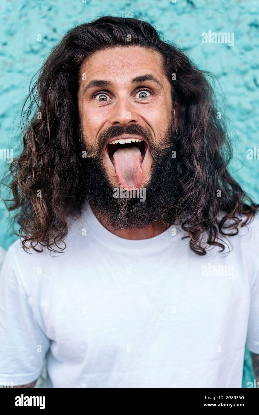 Cool bearded man with long hair sticking his tongue out and looking at  camera against blue wall Stock Photo - Alamy