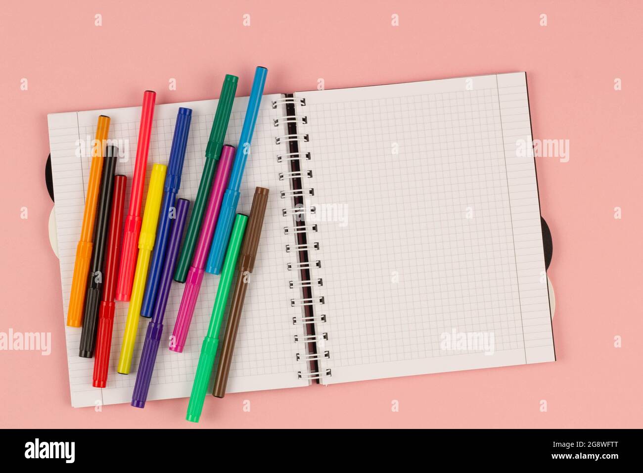 Open school squared notebook and colorful markers on the pink background. Blank white sheet of paper book on the table. Office supplies on the desktop Stock Photo