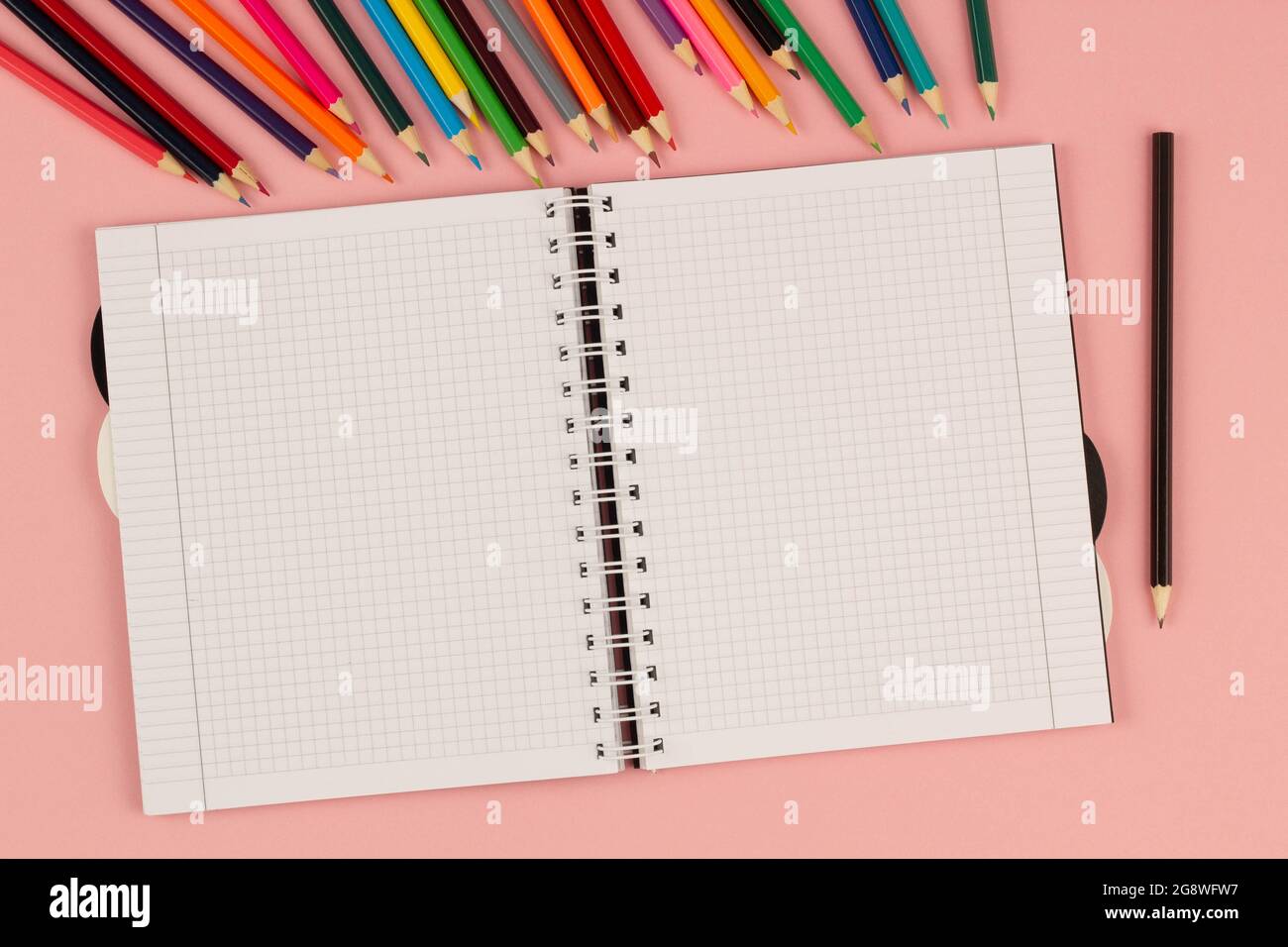 Open school squared notebook and colorful pencils on the pink background. Blank white sheet of paper book on the table. Office supplies on the desktop Stock Photo
