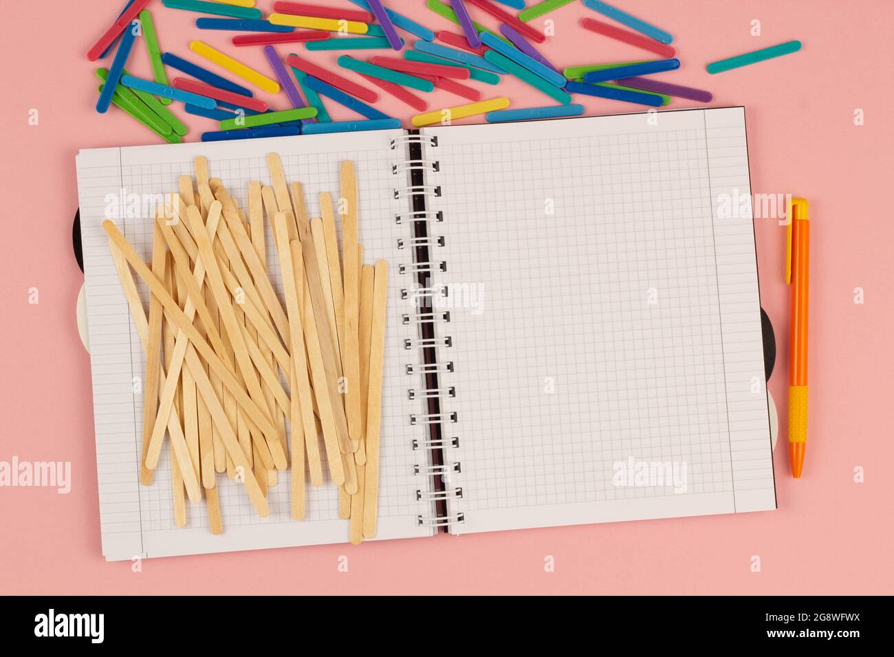 Open school squared notebook, pen, popsicle, colorful math sticks on the pink background. Blank white sheet of paper book on the table. Office Stock Photo