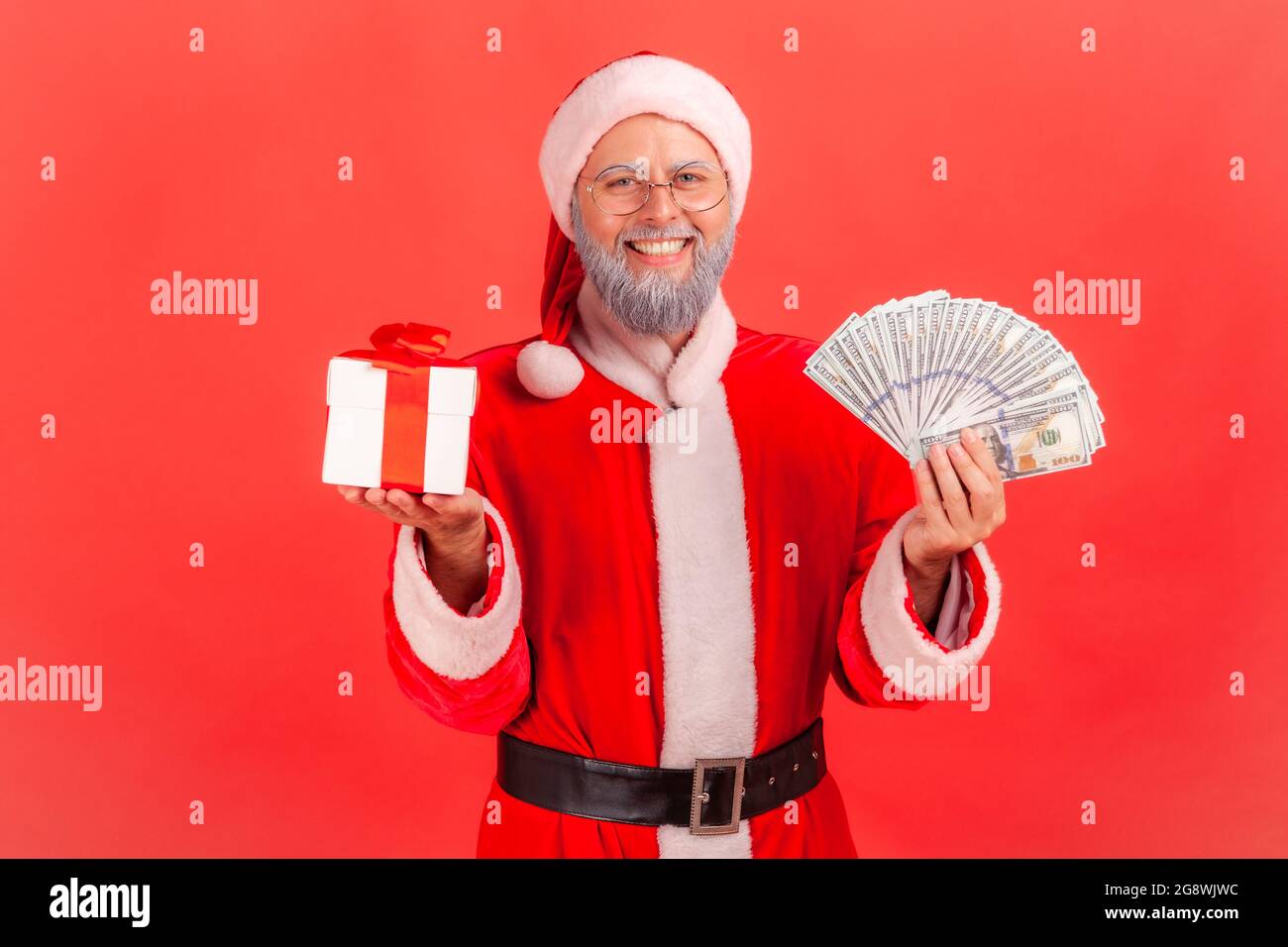Smiling elderly man with gray beard wearing santa claus costume holding fan of money and wrapped present box, looking at camera with toothy smile. Ind Stock Photo