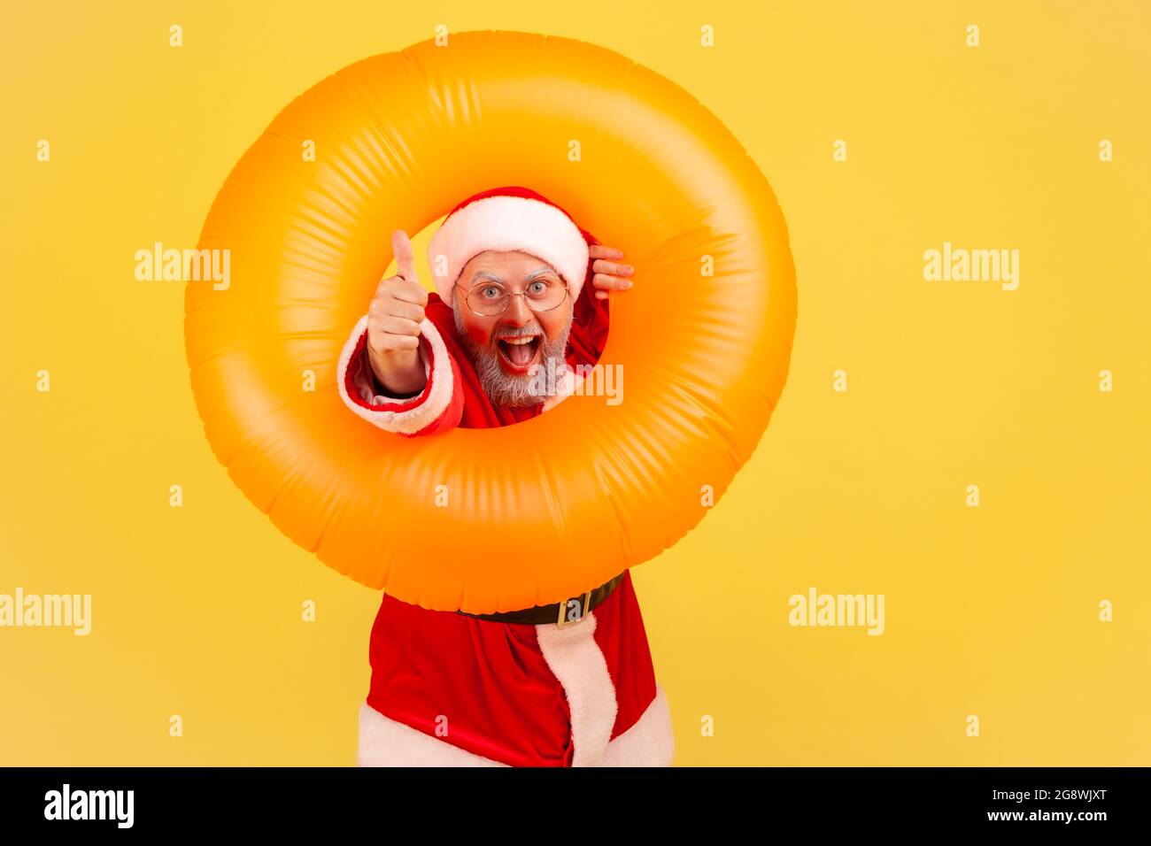 Extremely happy elderly man with gray beard wearing santa claus costume posing with orange rubber ring and showing thumb up, enjoying winter vacation. Stock Photo