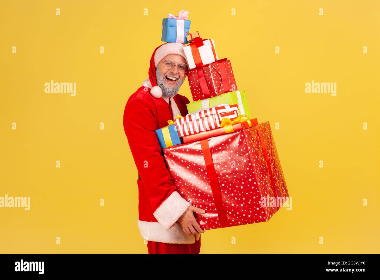 Happy funny elderly man with gray beard in santa claus costume holding stack of Christmas presents in hands and on head, celebrating with new year. In Stock Photo