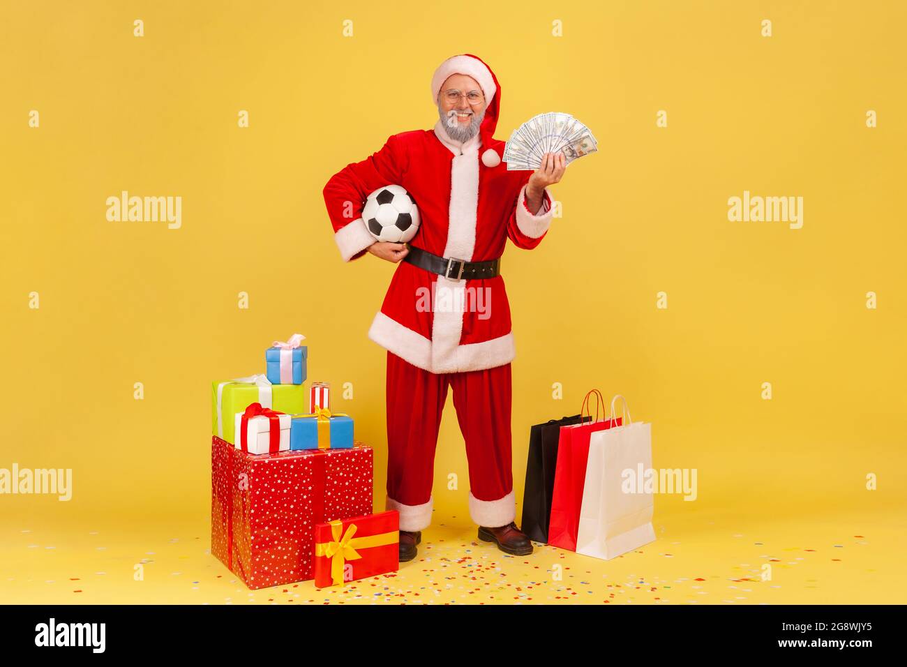 Happy satisfied elderly man wearing santa claus costume standing holding soccer ball and fan of money, betting and win, buy presents for Christmas. In Stock Photo
