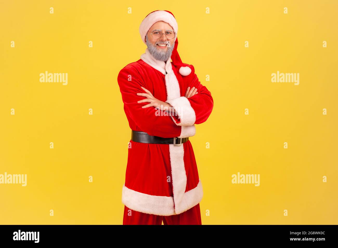 Positive smiling elderly man with gray beard wearing santa claus costume looking directly at camera, keeping hands folded, happy confident expression. Stock Photo