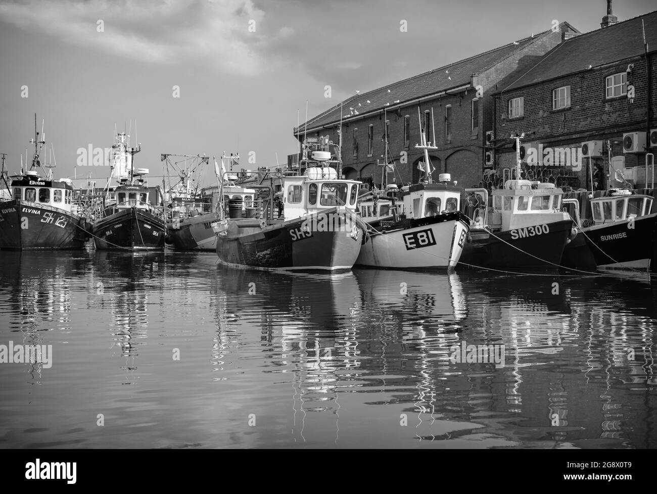 The harbour at Scarborough.  Fishing boats are moored in the harbour alongside a wharf with red-brick buildings.  A cloudy sky is above. Stock Photo