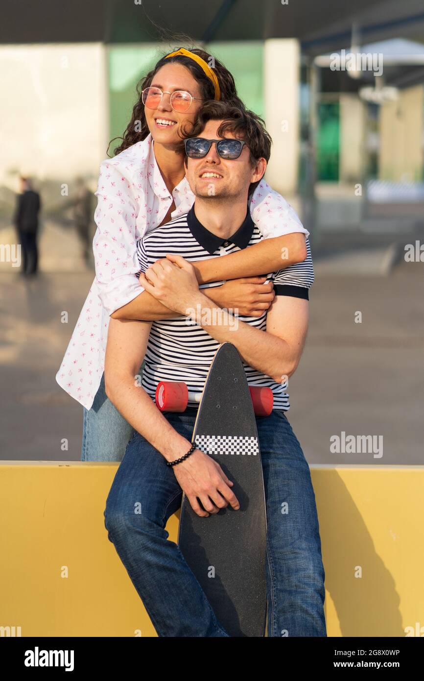 Young couple rest after longboarding: happy male and female in sunglasses enjoy sunset sit cuddling Stock Photo