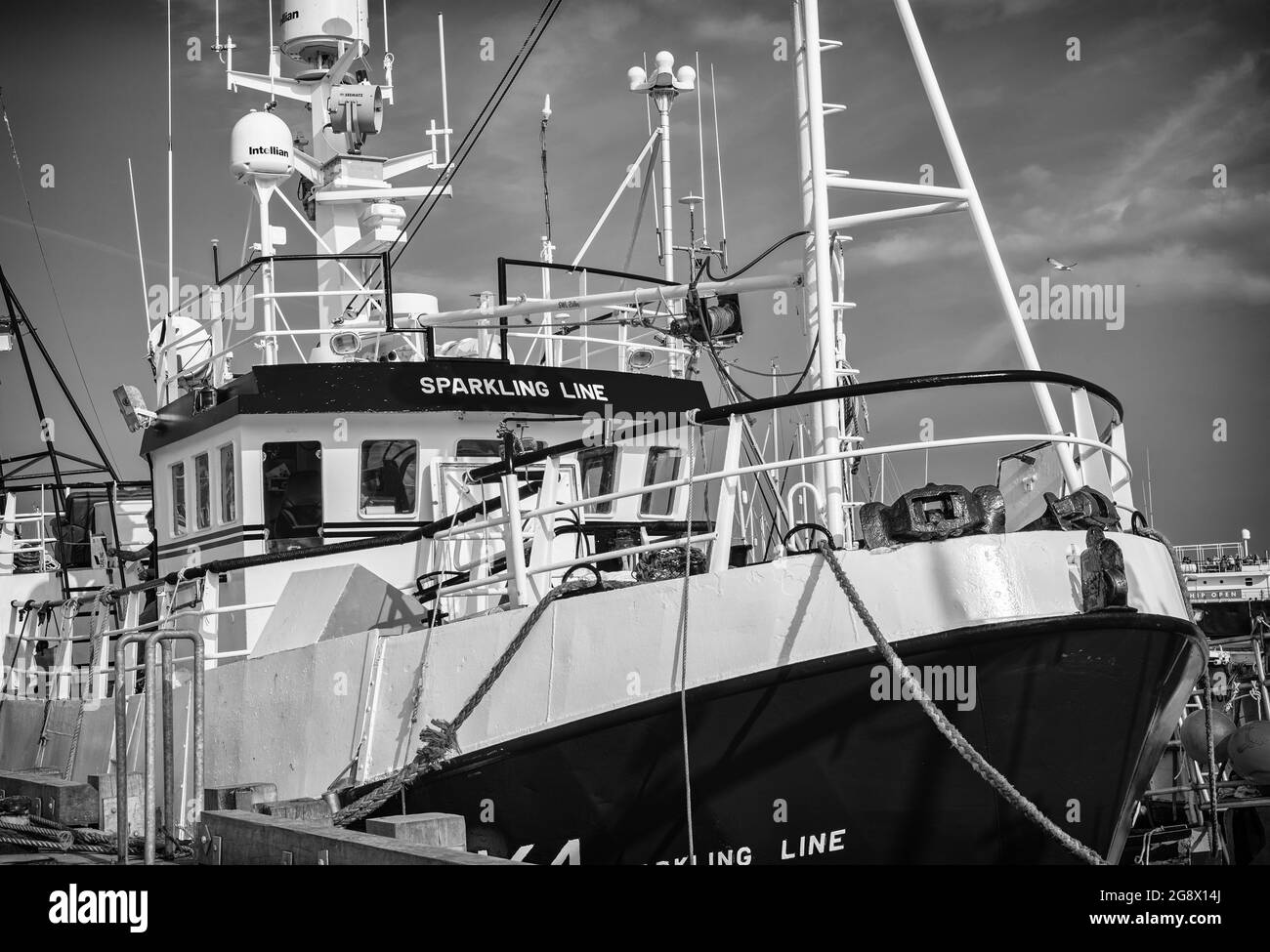 A freshly painted trawler is moored alongside a wharf.  Antennae and radar bristle above the bridge and a sky with clouds is above. Stock Photo